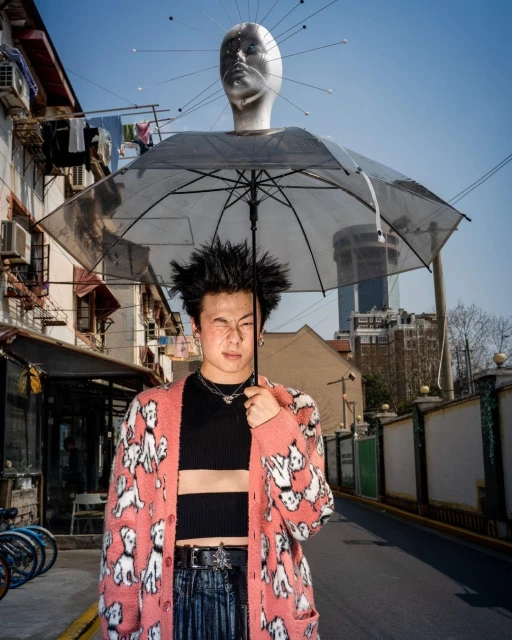 A young adult in a patterned cardigan stands on a street under an umbrella with a mannequin head hovering above him, creating a surreal scene.