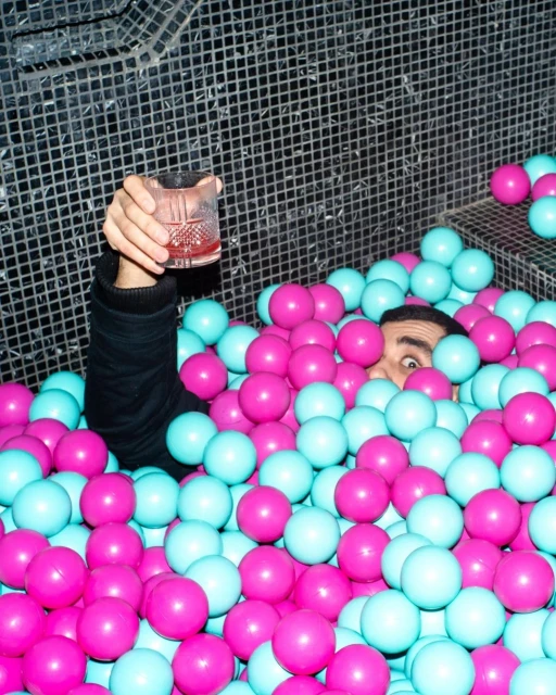 Adult man in a ball pit holding a cocktail glass filled with red liquid, dark hair, and a black top, indoor setting, and mosaic wall.