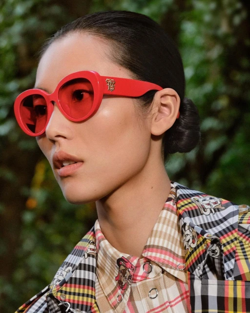 Woman with red sunglasses in a close-up shot. She's wearing a patterned shirt and has her dark hair pulled back, set against a green backdrop.