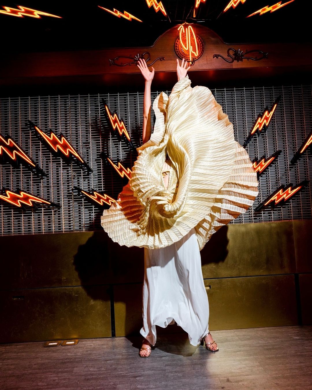 Adult female wearing a couture pleated dress with arms raised in an indoor environment with neon lightning bolt signs on the walls.