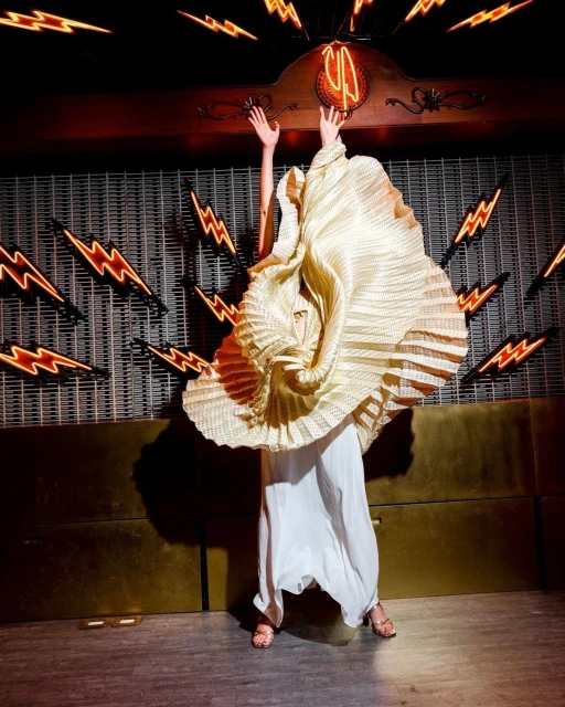 Adult female wearing a couture pleated dress with arms raised in an indoor environment with neon lightning bolt signs on the walls.