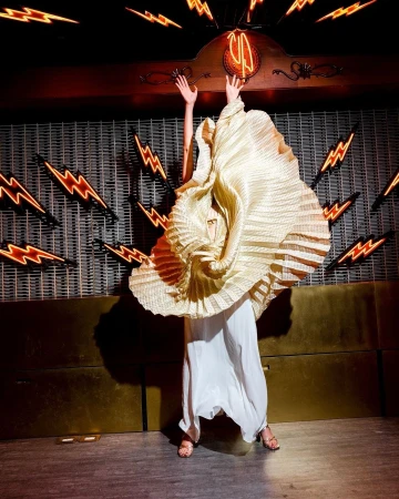 Adult female wearing a couture pleated dress with arms raised in an indoor environment with neon lightning bolt signs on the walls.