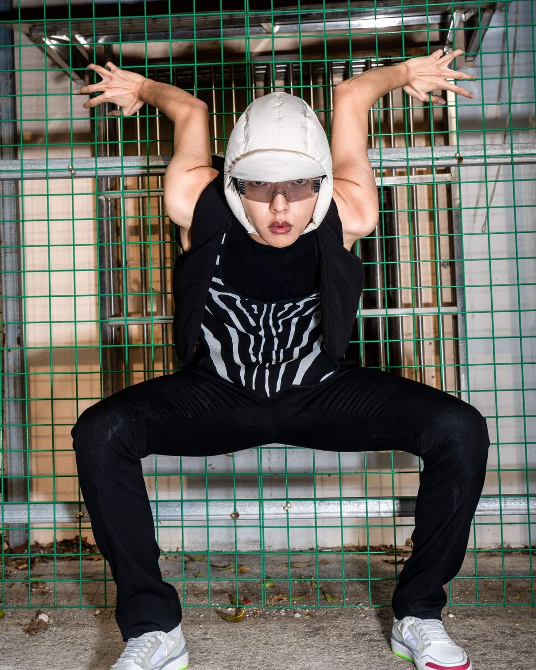 Person crouches behind green metal fence in a zebra-striped top, black pants, white helmet and sneakers with outstretched arms.