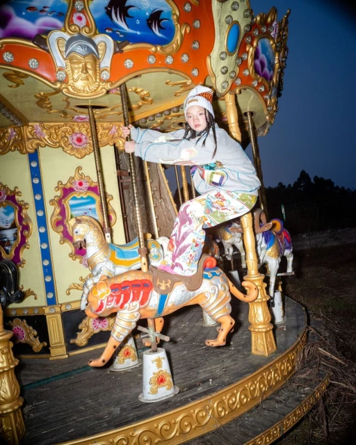 A young woman wearing a patterned outfit rides an animal figure on a vintage carousel, with trees and a dark blue sky in the background.