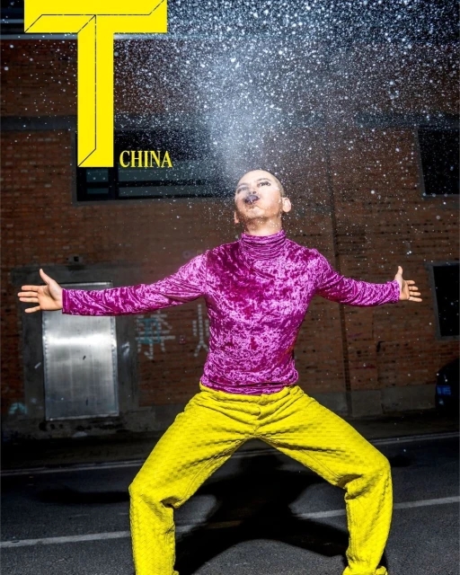 An adult male in a vibrant purple velvet top and patterned yellow pants stands on an asphalt street in front of a brick wall.
