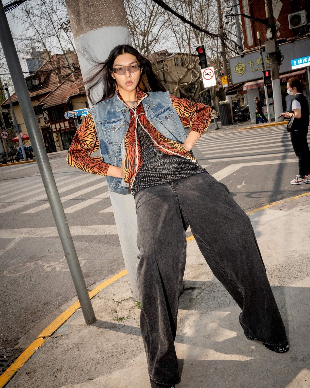 Model wearing a denim vest over a tiger-stripe shirt and wide-leg black jeans leaning against a pole on a street.