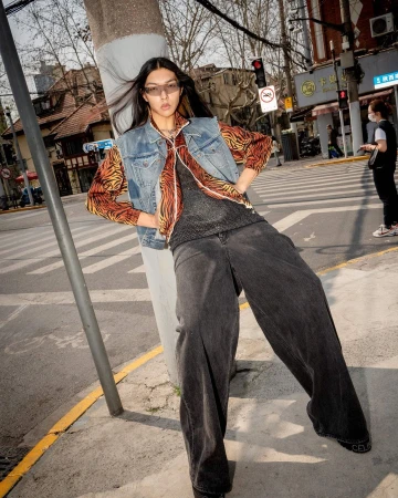 Model wearing a denim vest over a tiger-stripe shirt and wide-leg black jeans leaning against a pole on a street.