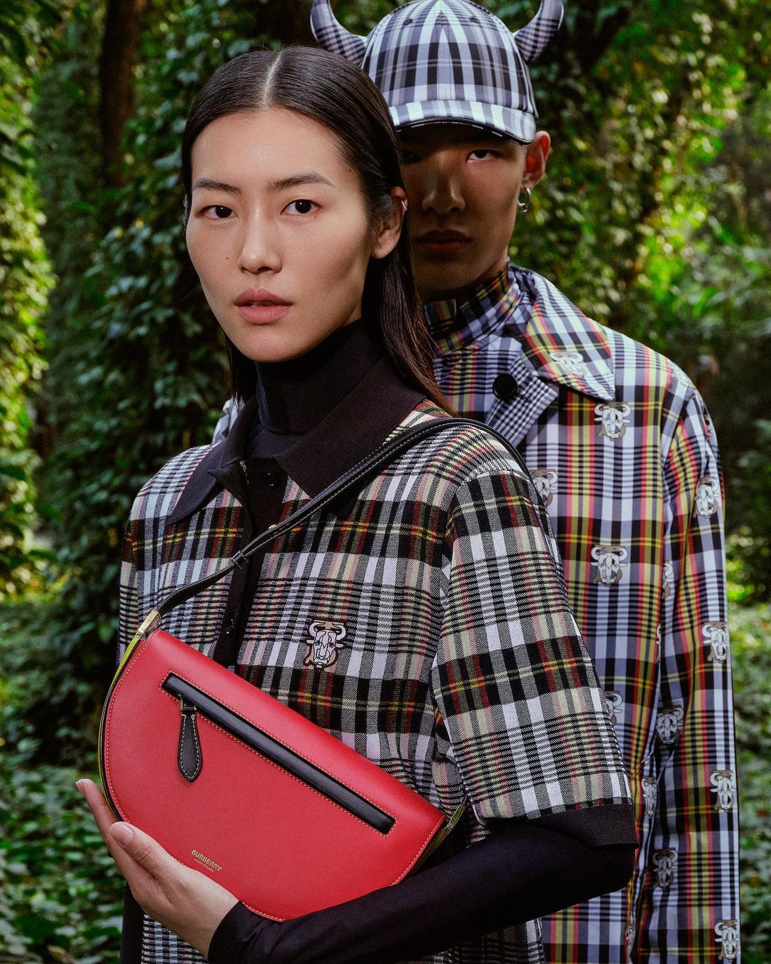 Two models showcase plaid Burberry clothing in a forest setting. An Asian woman holds a bright red purse, posing in the foreground.