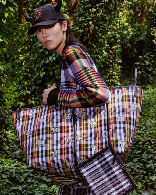 A young Asian model in an outdoor fashion shot wearing a plaid jacket, carrying a large Burberry plaid tote bag and wearing a horned cap.