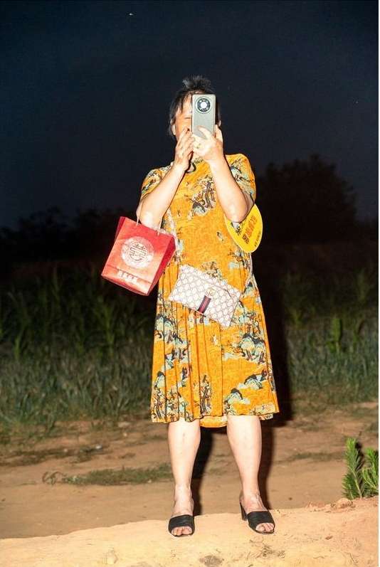Adult female standing outdoors at night, using a smartphone for photography, with handbag, gift bag, wearing an orange dress.