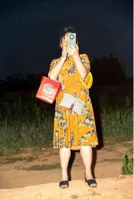 Adult female standing outdoors at night, using a smartphone for photography, with handbag, gift bag, wearing an orange dress.