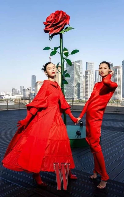 Two women in red outfits pose near a giant rose sculpture on a rooftop with city skyline backdrop, under a clear blue sky.