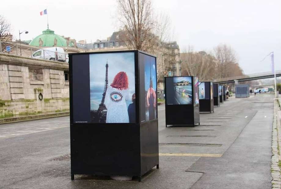 Outdoor art installation in Paris features photos of models with the Eiffel Tower as backdrop on a cloudy day.