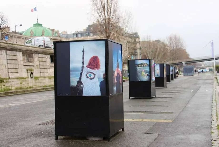 Outdoor art installation in Paris features photos of models with the Eiffel Tower as backdrop on a cloudy day.