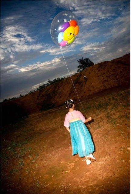 A girl in a blue dress holds a smiley-face balloon outdoors, with a dirt landscape and a cloudy blue sky in the background.