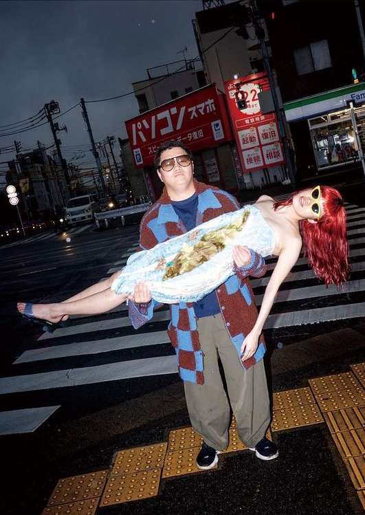 Man carries woman across a crosswalk at night. The man is wearing a sweater and glasses, and the woman is in a light dress.