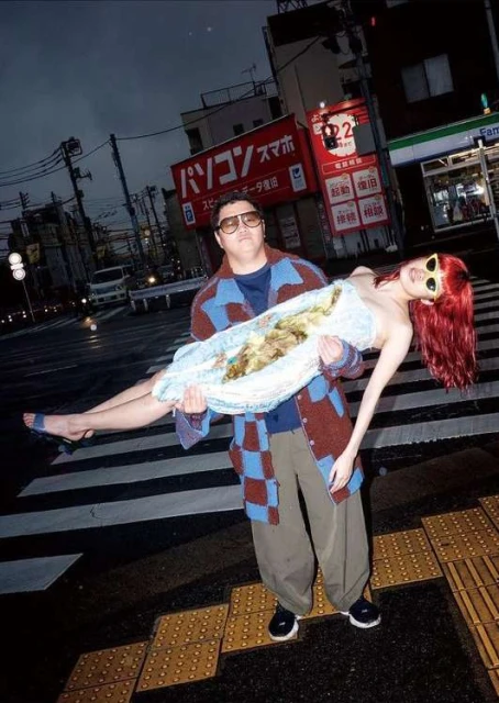 Man carries woman across a crosswalk at night. The man is wearing a sweater and glasses, and the woman is in a light dress.