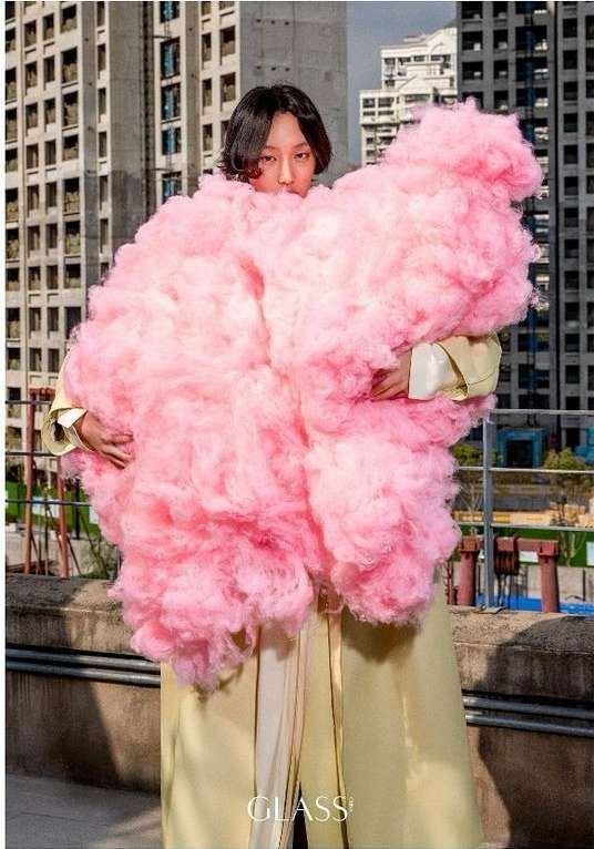 Adult poses outdoors wearing a large, pink, cloud-like tulle sculpture with beige, satin-like wide leg pants near buildings.