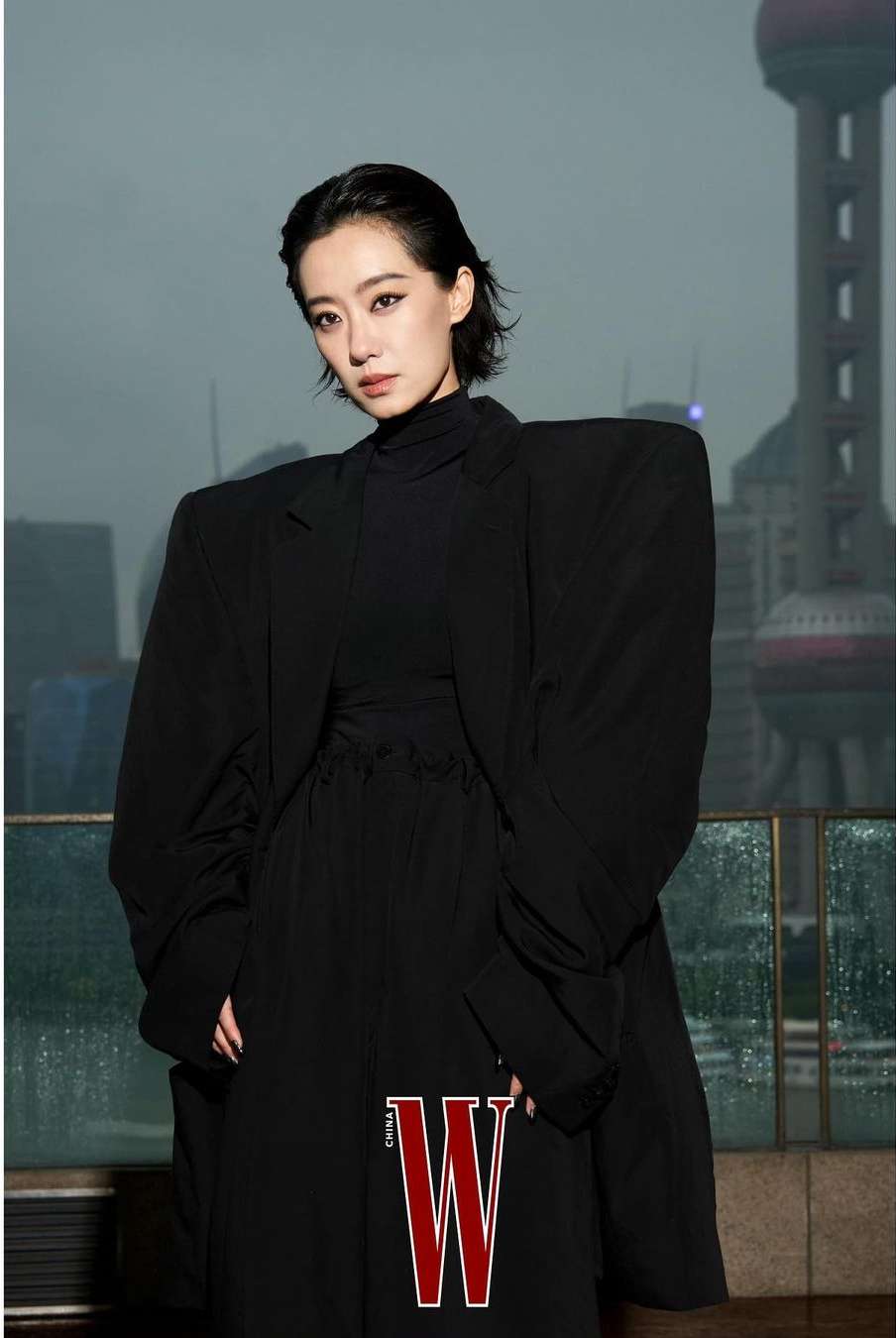 Woman in oversized black blazer poses in front of city skyline with W magazine logo visible. Fashion editorial portrait.