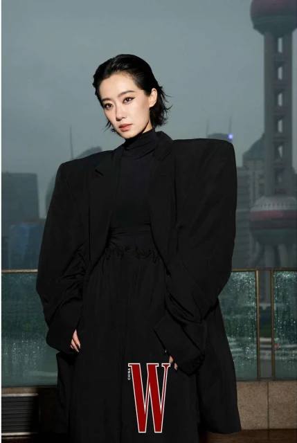 Woman in oversized black blazer poses in front of city skyline with W magazine logo visible. Fashion editorial portrait.