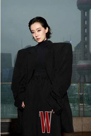 Woman in oversized black blazer poses in front of city skyline with W magazine logo visible. Fashion editorial portrait.