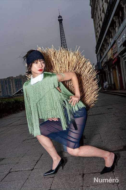 Editorial fashion shot of a woman in a green fringe top and sheer skirt kneeling on a stone street in an urban setting with the Eiffel Tower.