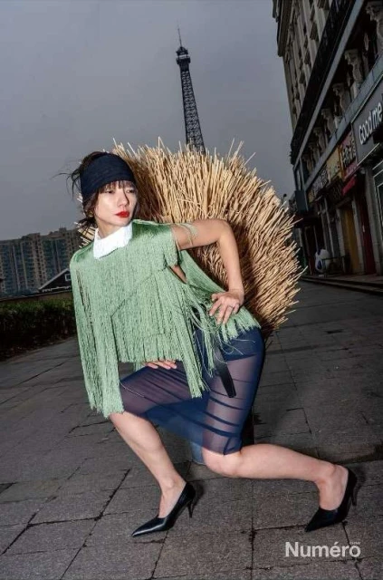 Editorial fashion shot of a woman in a green fringe top and sheer skirt kneeling on a stone street in an urban setting with the Eiffel Tower.
