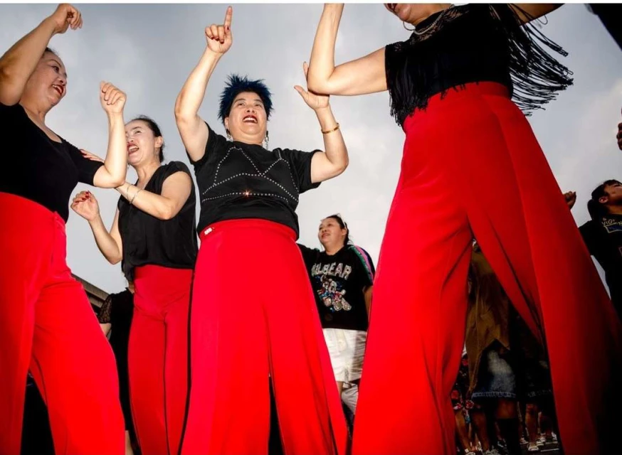 A group of Asian women in red pants and black shirts dancing outdoors with excited expressions in a low-angle shot against a cloudy sky.