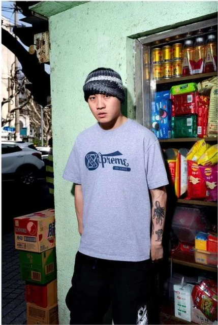 An adult male stands outdoors wearing a grey Supreme tee, black pants, and a beanie, with a convenience store shelf to his side.