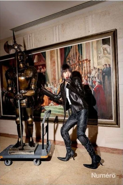 A fashion shot of an adult female in black leather posing next to a knight statue in front of an elaborate painting in an art gallery.