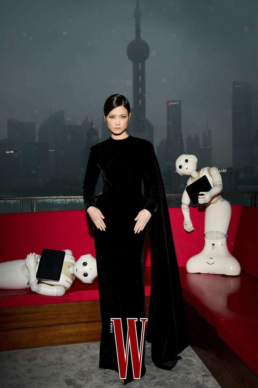 A woman in a black gown stands near two robots on a red couch, with the Shanghai skyline in the background, for a fashion editorial.
