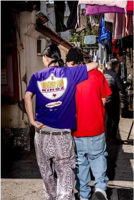 Two people wearing graphic T-shirts and jeans walk on a street lined with clotheslines in an urban alley with gray walls and pavement.