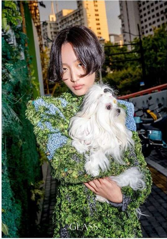 Asian woman holding a fluffy white dog in an urban setting, wearing a green textured coat and standing near buildings, bathed in soft light.
