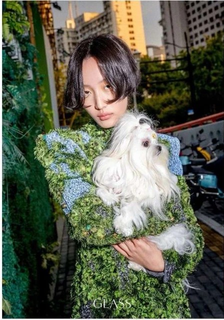 Asian woman holding a fluffy white dog in an urban setting, wearing a green textured coat and standing near buildings, bathed in soft light.