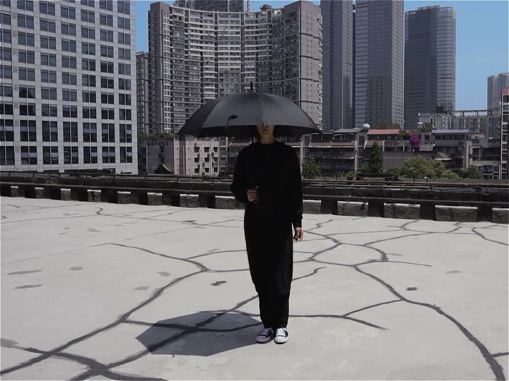 Adult female standing outdoors on cracked concrete in a city environment, wearing all black and holding an umbrella over her head.