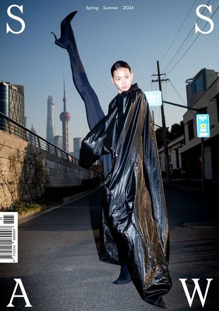 Editorial fashion shot of a woman in a black vinyl robe posing on a street with an elevated leg in a cityscape backdrop.