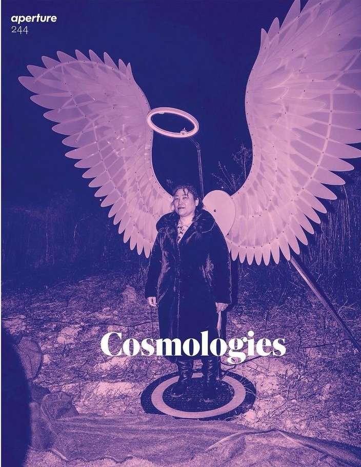 Magazine cover with an adult female standing wearing angel wings, ring light halo, dark coat, and boots in a nighttime outdoor environment.