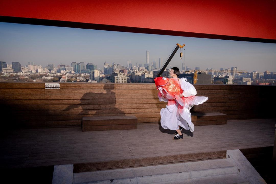 Person in a large goldfish costume standing on a rooftop with a cityscape in the background, under a red overhang.