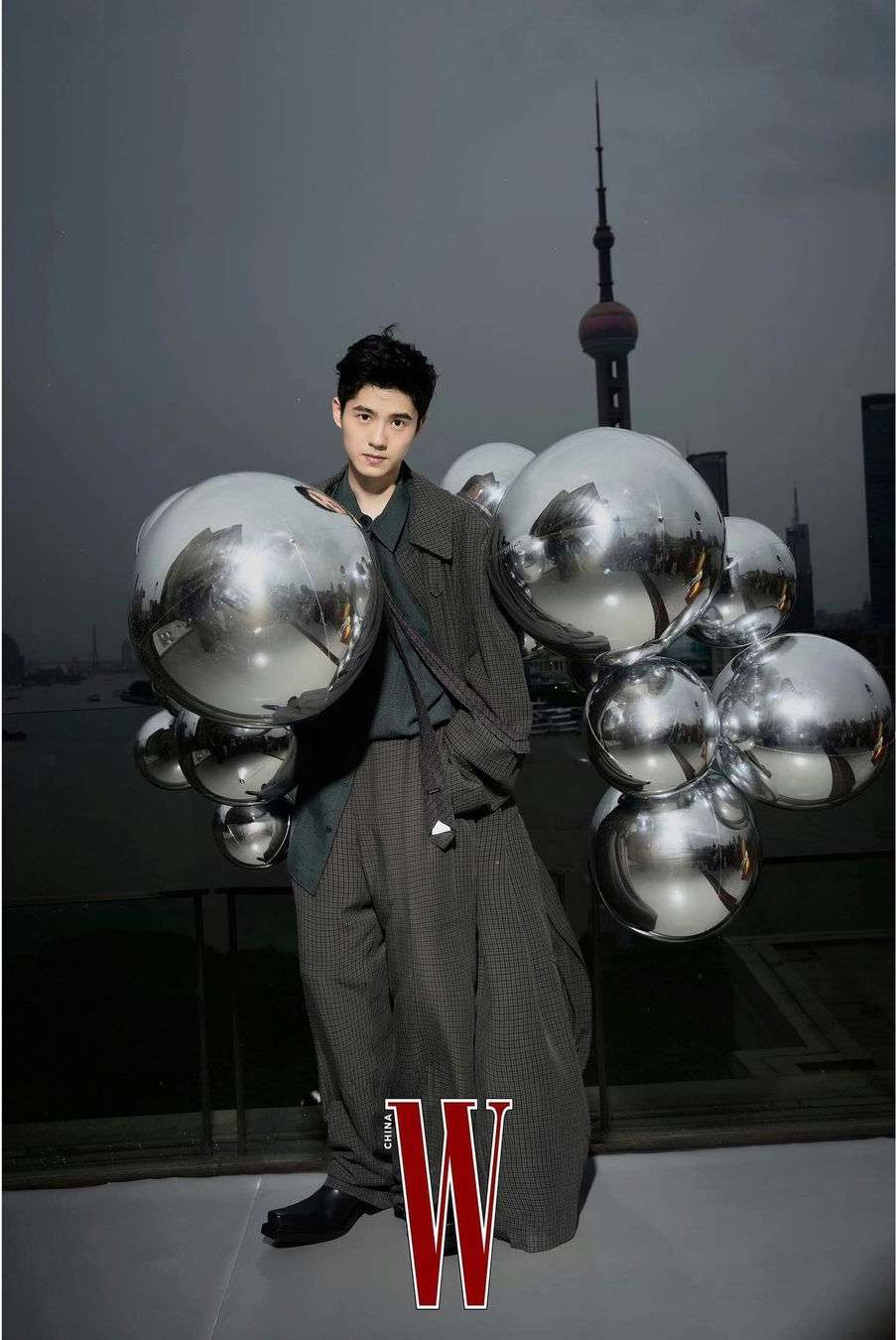 Portrait of a young Asian man in a stylish grey outfit posing with reflective spheres against a Shanghai cityscape backdrop; fashion editorial.