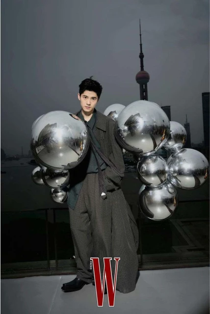 Portrait of a young Asian man in a stylish grey outfit posing with reflective spheres against a Shanghai cityscape backdrop; fashion editorial.