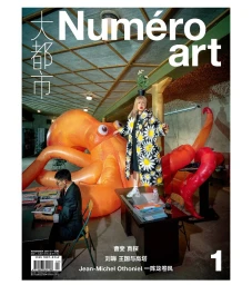 A woman in a floral coat standing on a desk beside a giant inflatable orange octopus, with a seated man reading nearby.