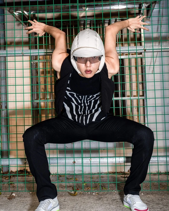 Model crouching with arms raised against a green wire fence wearing a white padded cap and zebra print top