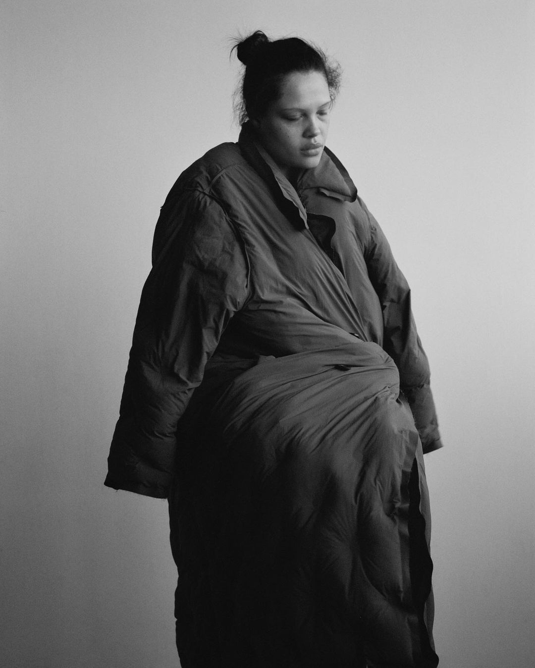 Monochrome portrait of a woman with a bun, wearing an oversized puffer coat, standing against a light background, creating a fashion editorial image.