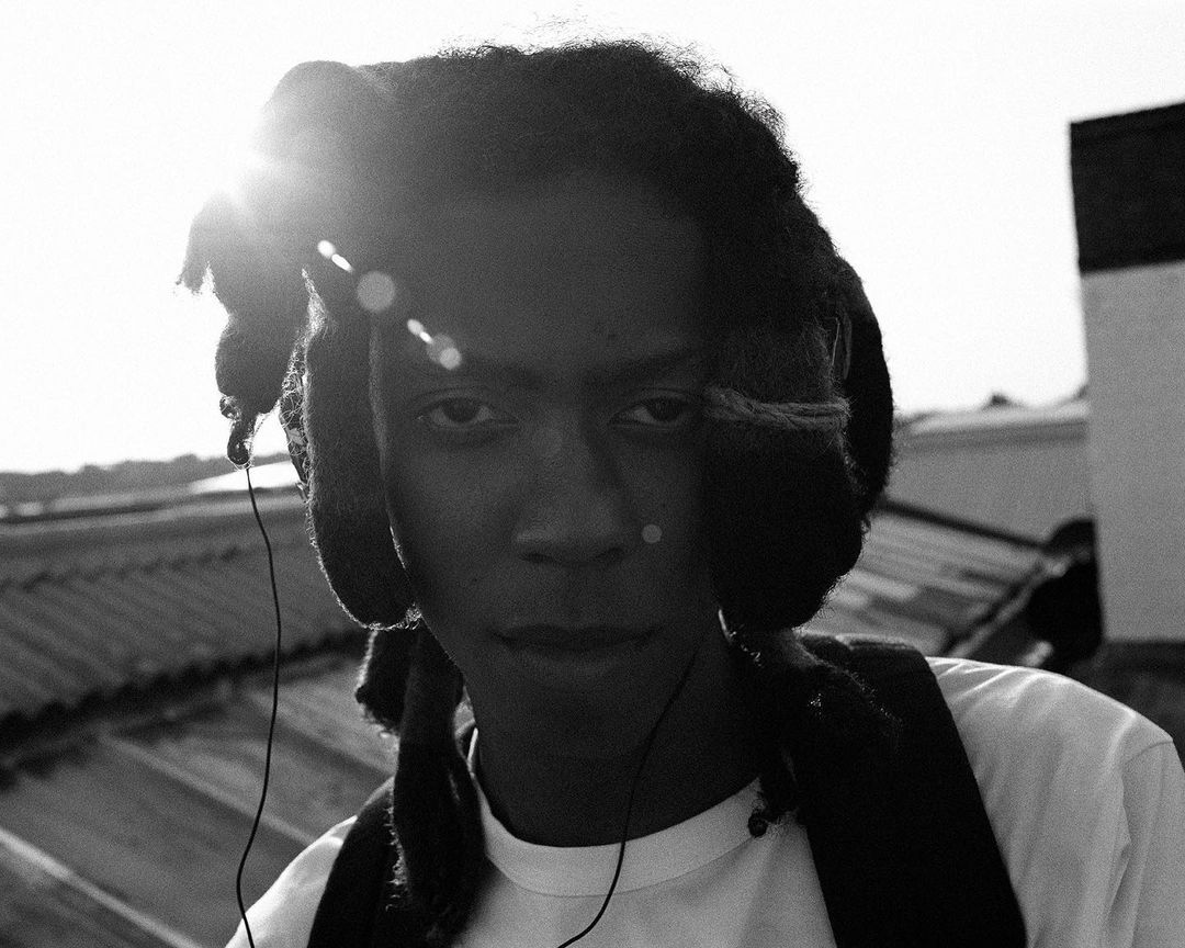 Monochrome portrait of an adult male wearing headphones and a backpack, with sunlight creating lens flare against a rooftop background.