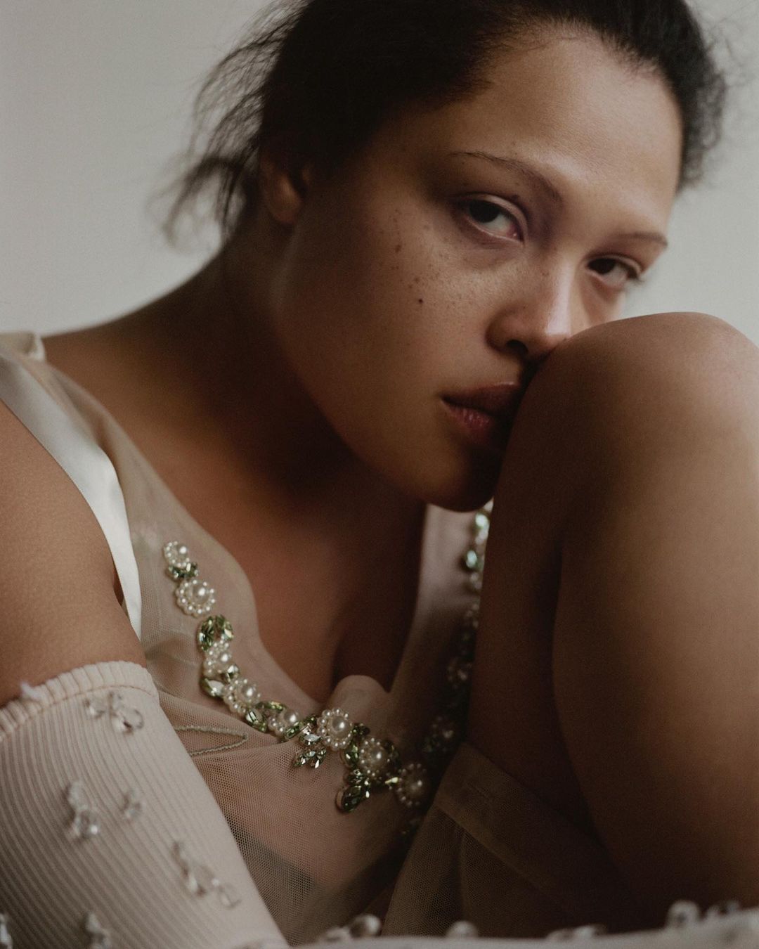 Portrait of a woman with light brown hair and embellished dress, resting her cheek on her knee in a soft, indoor setting.