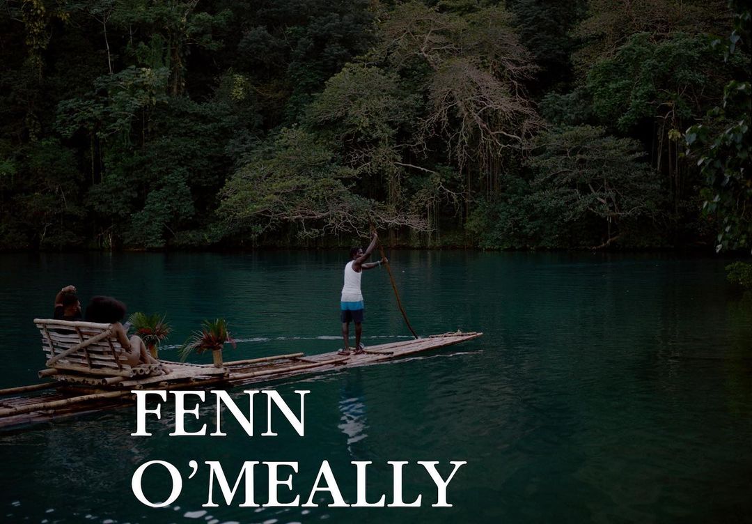 Two people ride on a bamboo raft being poled by a man in a river surrounded by lush trees and vegetation in an outdoor setting.