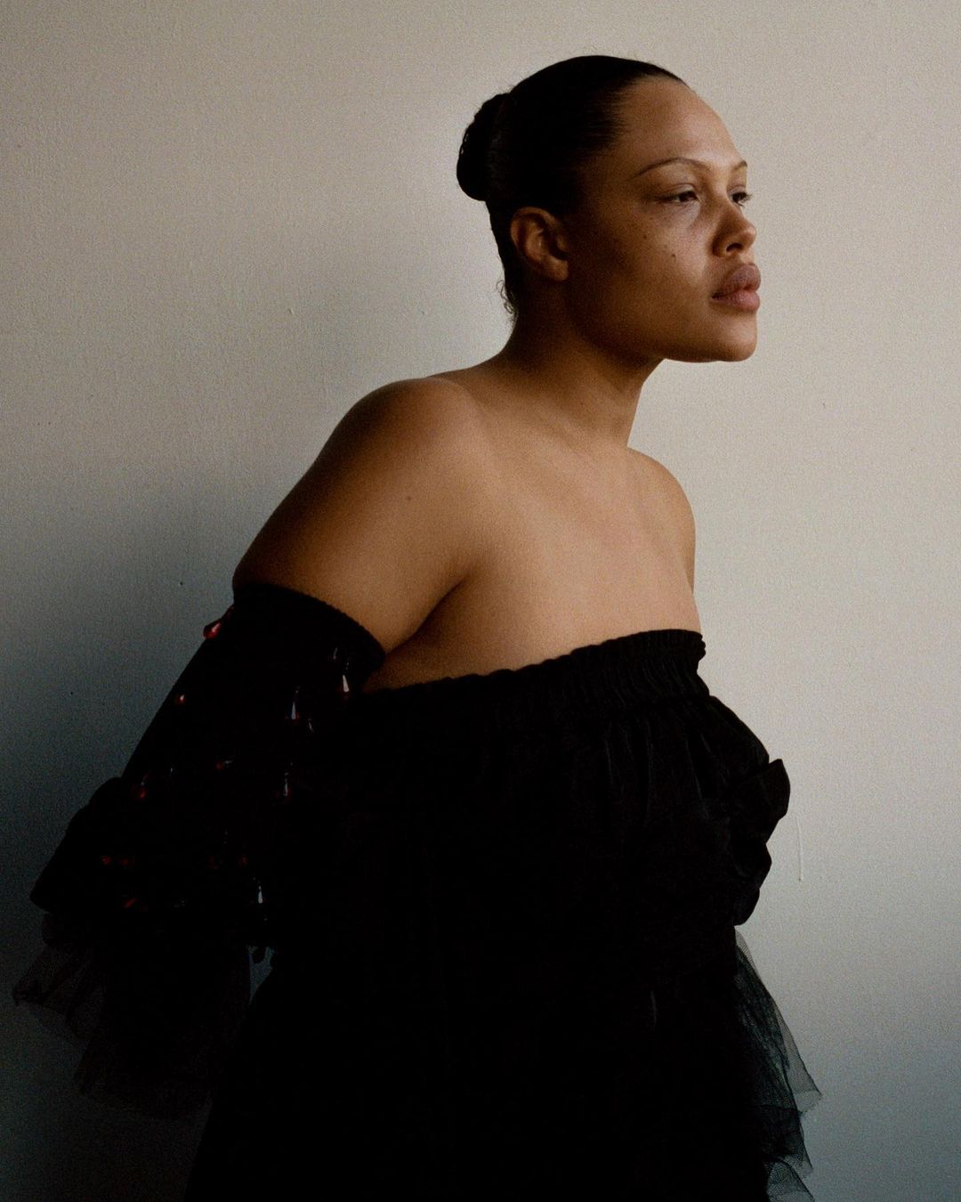 Elegant woman in a strapless black tulle dress with off-the-shoulder sleeves poses indoors against a plain wall in a side view.