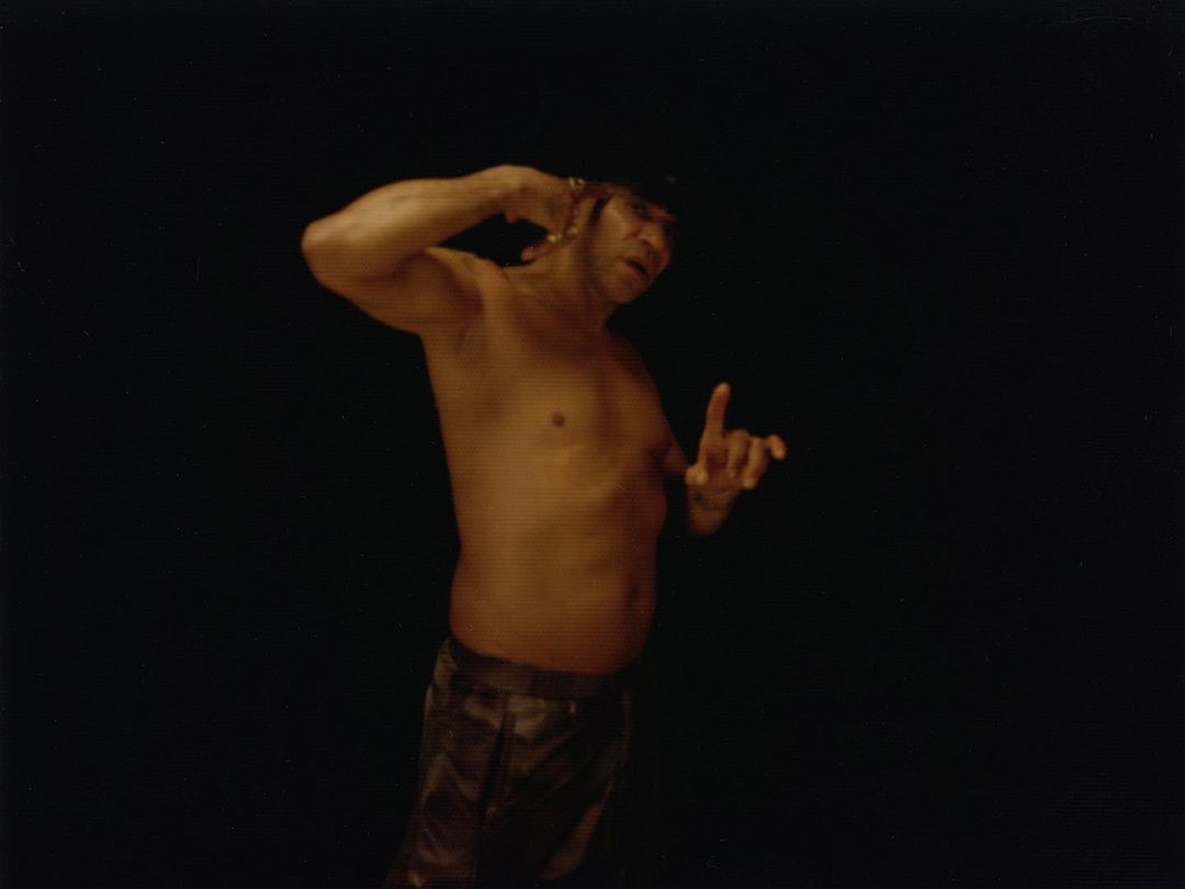 Shirtless man with bandana stands in a dark setting with hand gestures, creating a dramatic and artistic image.