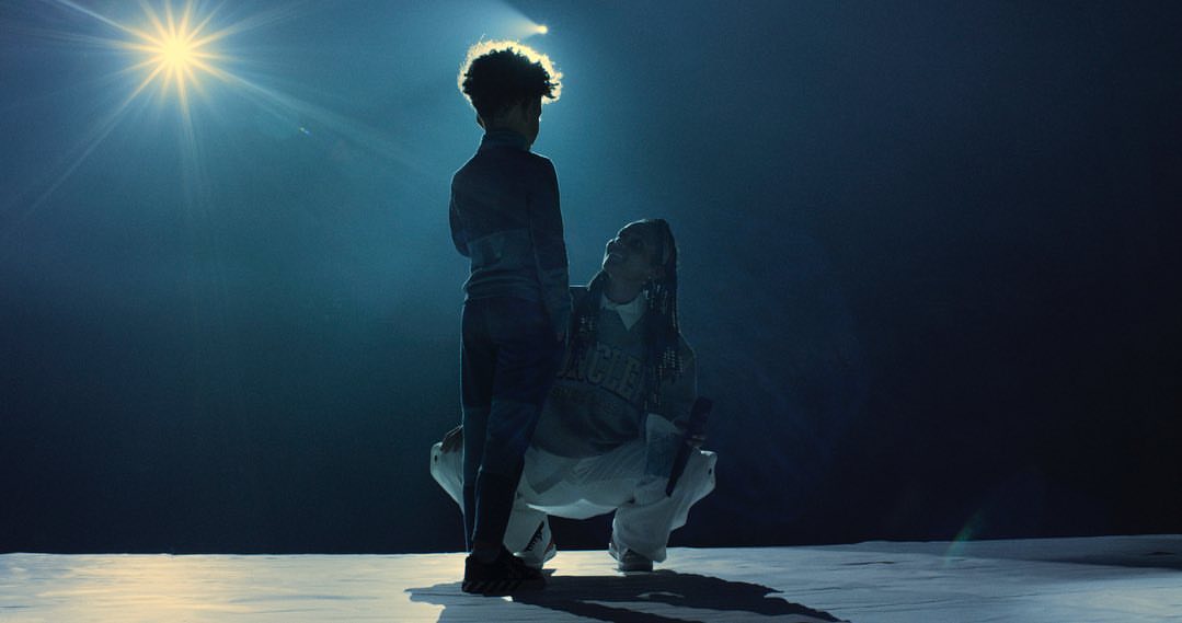 Two people, a child standing and an adult crouching, lit by a bright spotlight against a dark, monochromatic backdrop.