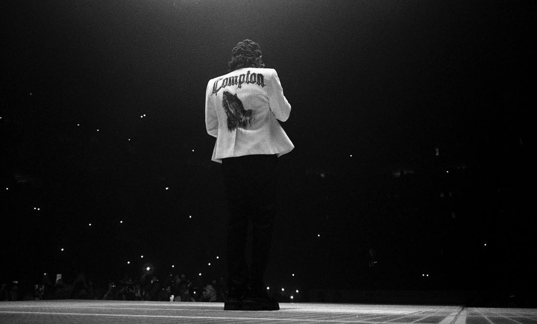 Male performer in white 'Compton' jacket on stage in low light, addressing a large crowd with cell phone flashlights visible.